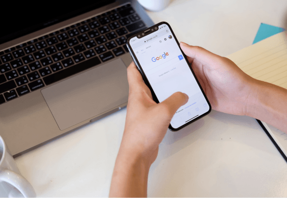 smartphone com tela mostrando buscador google e computador ao fundo